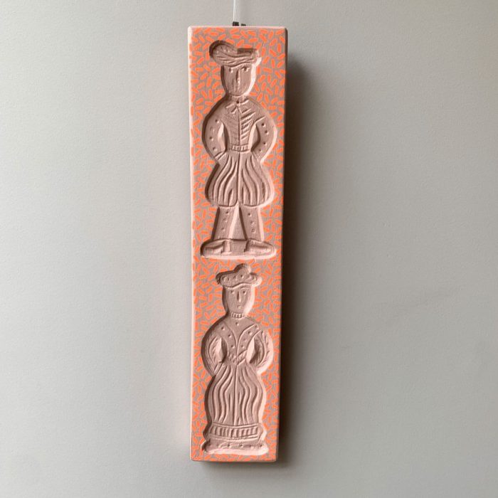 Opgehangen houten wanddecoratie aan een muur – met twee figuren in reliëf en fluorescerend oranje patroon op zachtroze achtergrond